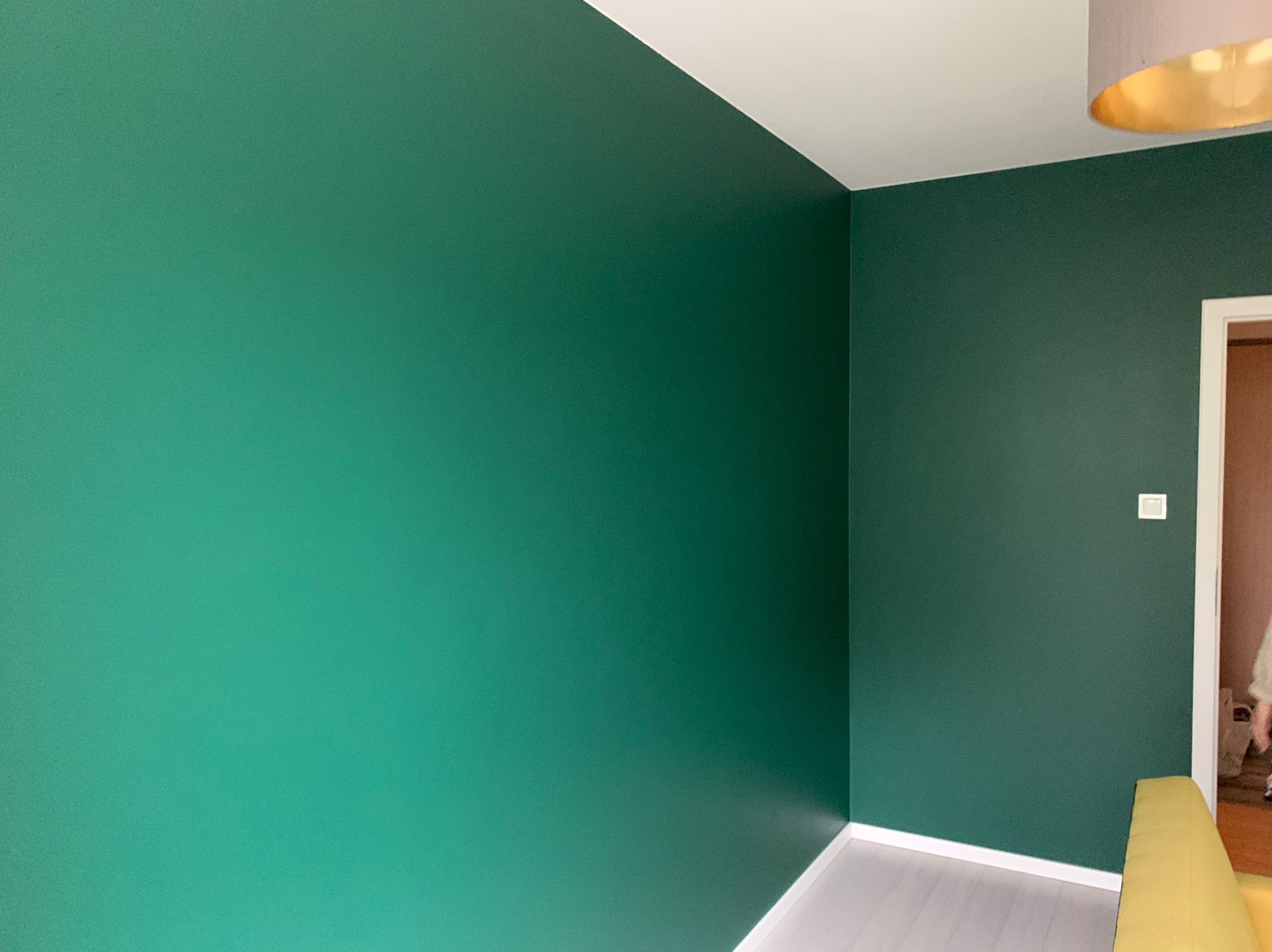 Ściana pomalowana na intensywny, butelkowy odcień zieleni w rogu pomieszczenia. Widoczny fragment lampy z złotym wnętrzem i białe listwy przypodłogowe.