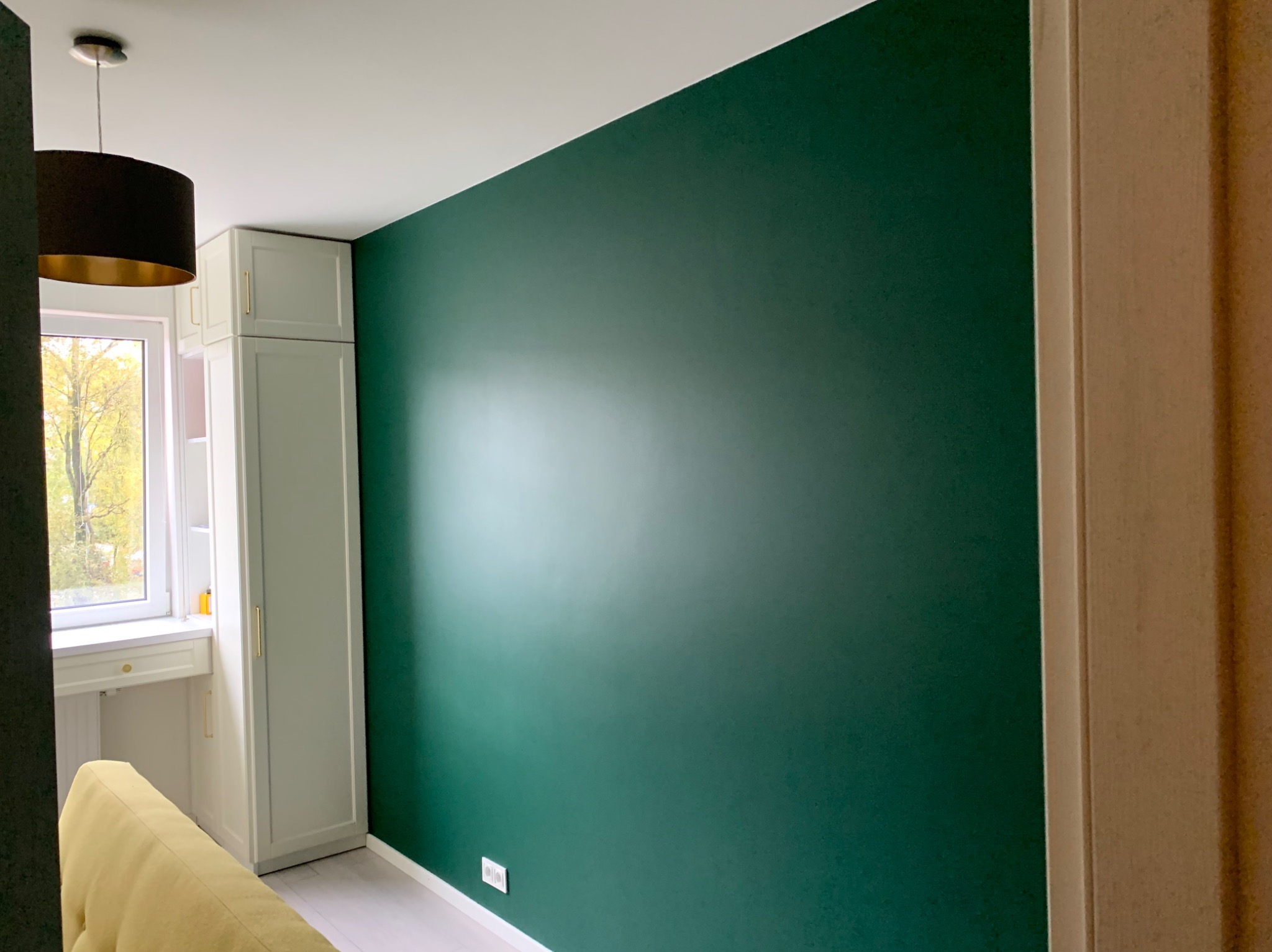 Ściana pomalowana na zielono w Luboniu, widoczne gniazdko elektryczne i fragment mebli. Gładka faktura farby, równomierne pokrycie. Nowoczesne wykończenie wnętrza.