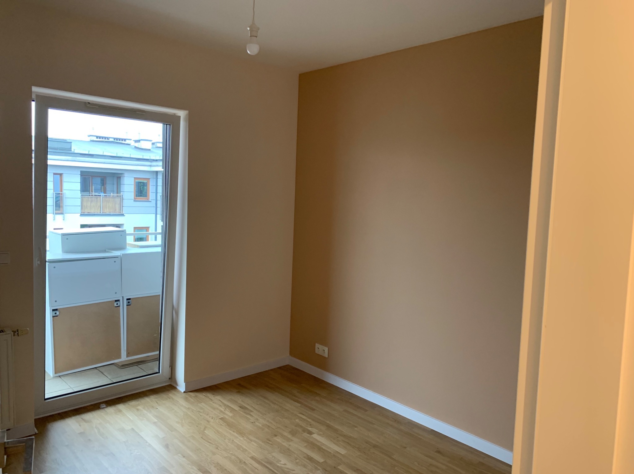 Pomalowane na beżowo wnętrze pokoju z drewnianą podłogą i balkonem z widokiem na sąsiedni budynek. Widoczne gniazdka elektryczne i białe listwy przypodłogowe.