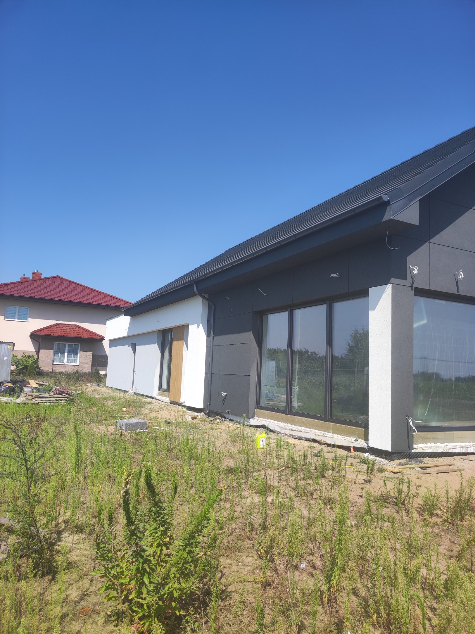 Nowoczesny dom z grafitową elewacją i dużymi oknami, w tle drugi dom z czerwoną dachówką. Widoczne prace wykończeniowe i niezagospodarowany teren.