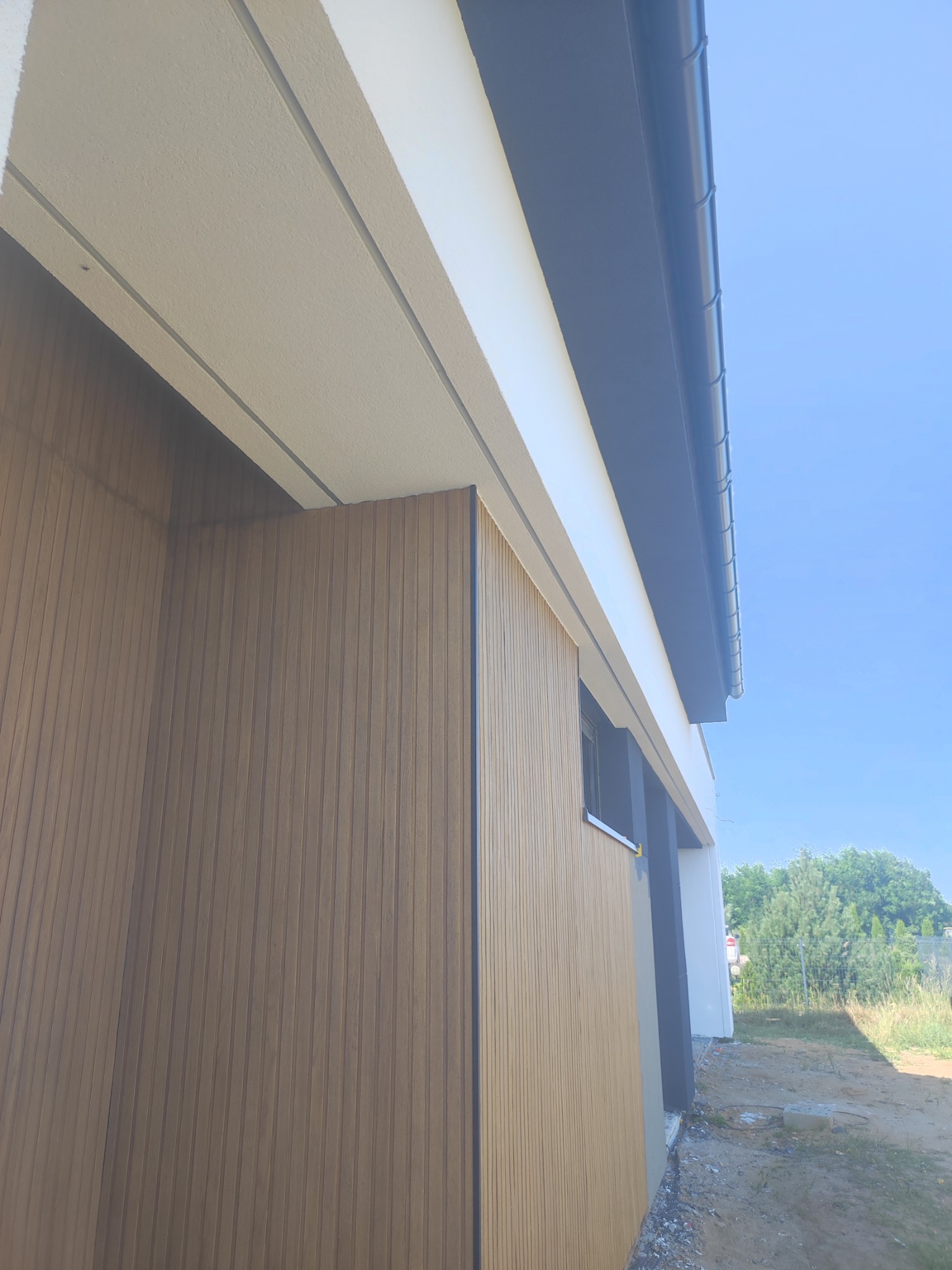 Nowoczesna elewacja domu z drewnianą okładziną w odcieniach brązu, widok z dołu, fragment okna i dachu, błękitne niebo w tle.