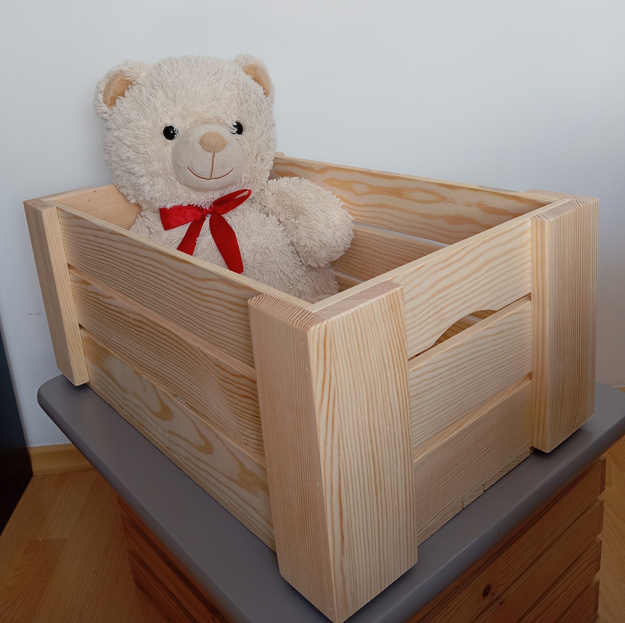 Drewniana skrzynka, ręcznie wykonana z jasnego drewna, z pluszowym misiem w środku. Widoczne detale stolarskie i naturalne usłojenie drewna.