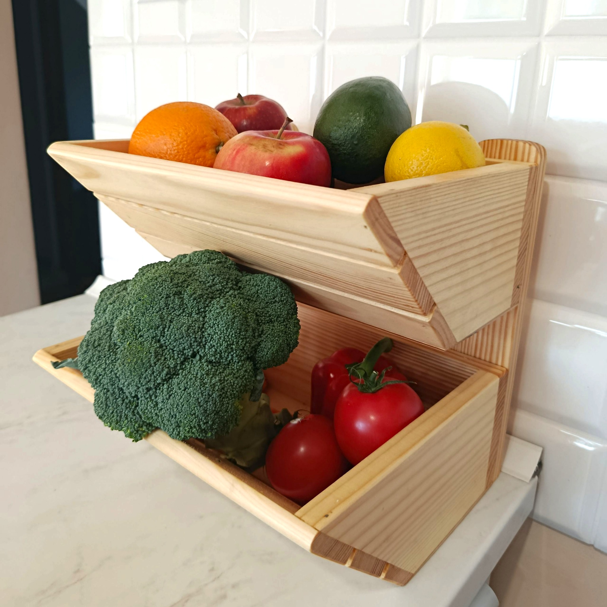 Dwupoziomowy, drewniany regał na owoce i warzywa. Na górze jabłka, pomarańcza, awokado i cytryna. Na dole brokuł, papryka i pomidory. Jasne drewno, białe tło.