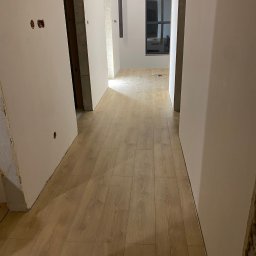 InProWnętrza - Korytarz w trakcie wykańczania: świeżo ułożone panele podłogowe, surowe ściany przygotowane do malowania, widoczne puszki elektryczne. W tle okno.