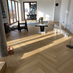 InProWnętrza - Jasna, drewniana podłoga ułożona w jodełkę w przestronnym, jasnym pokoju. Widoczne narzędzia i materiały budowlane sugerują etap prac wykończeniowych.