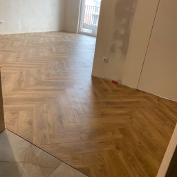 InProWnętrza - Ułożona podłoga z paneli w jodełkę w jasnym odcieniu drewna, widok z progu. Ściany w trakcie wykończenia, widoczne gniazdka i włączniki. Nowe panele podłogowe.