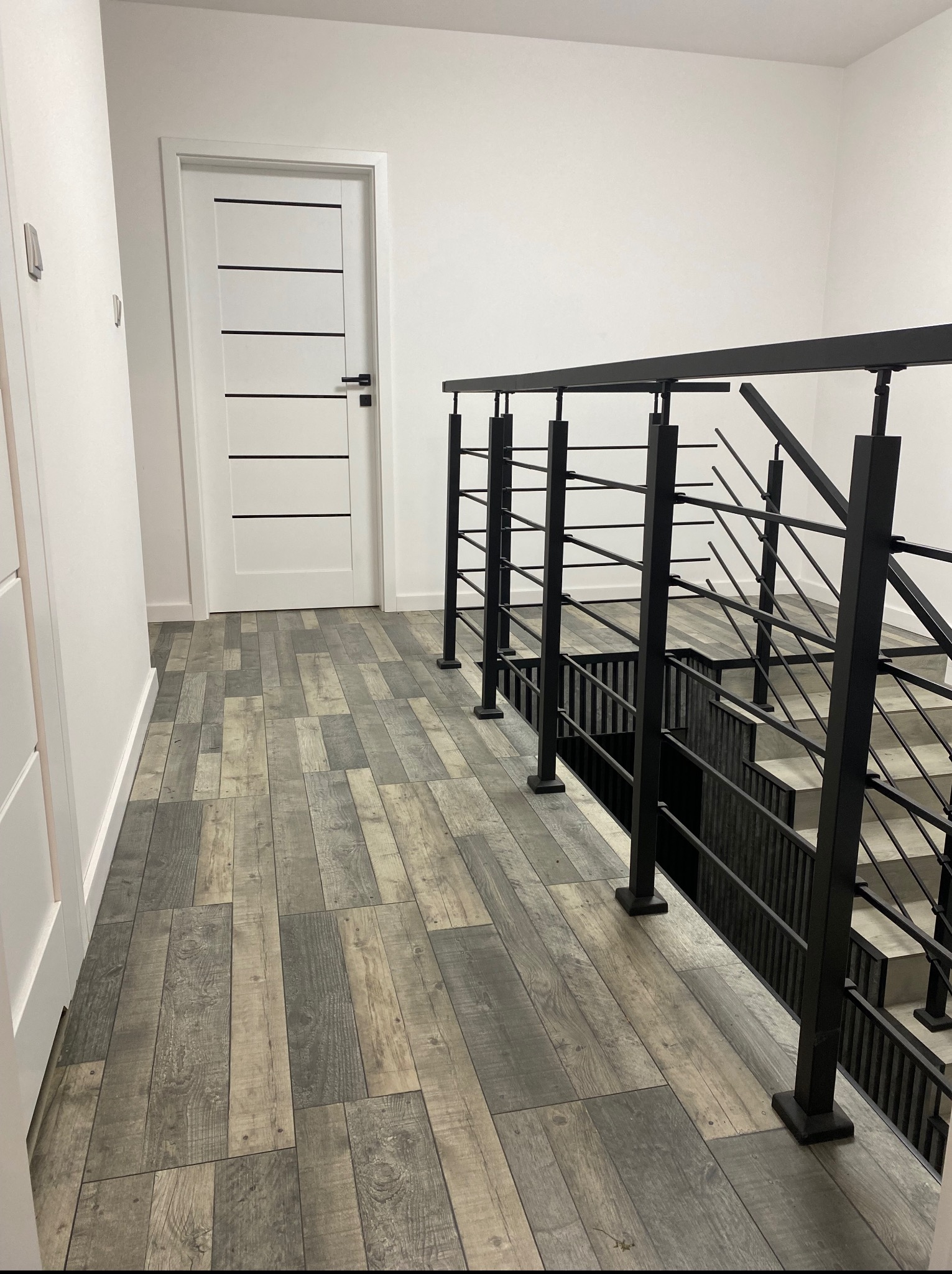 Nowoczesne wnętrze: szare panele podłogowe imitujące drewno, białe ściany i drzwi z czarnymi akcentami, czarna metalowa balustrada schodowa.