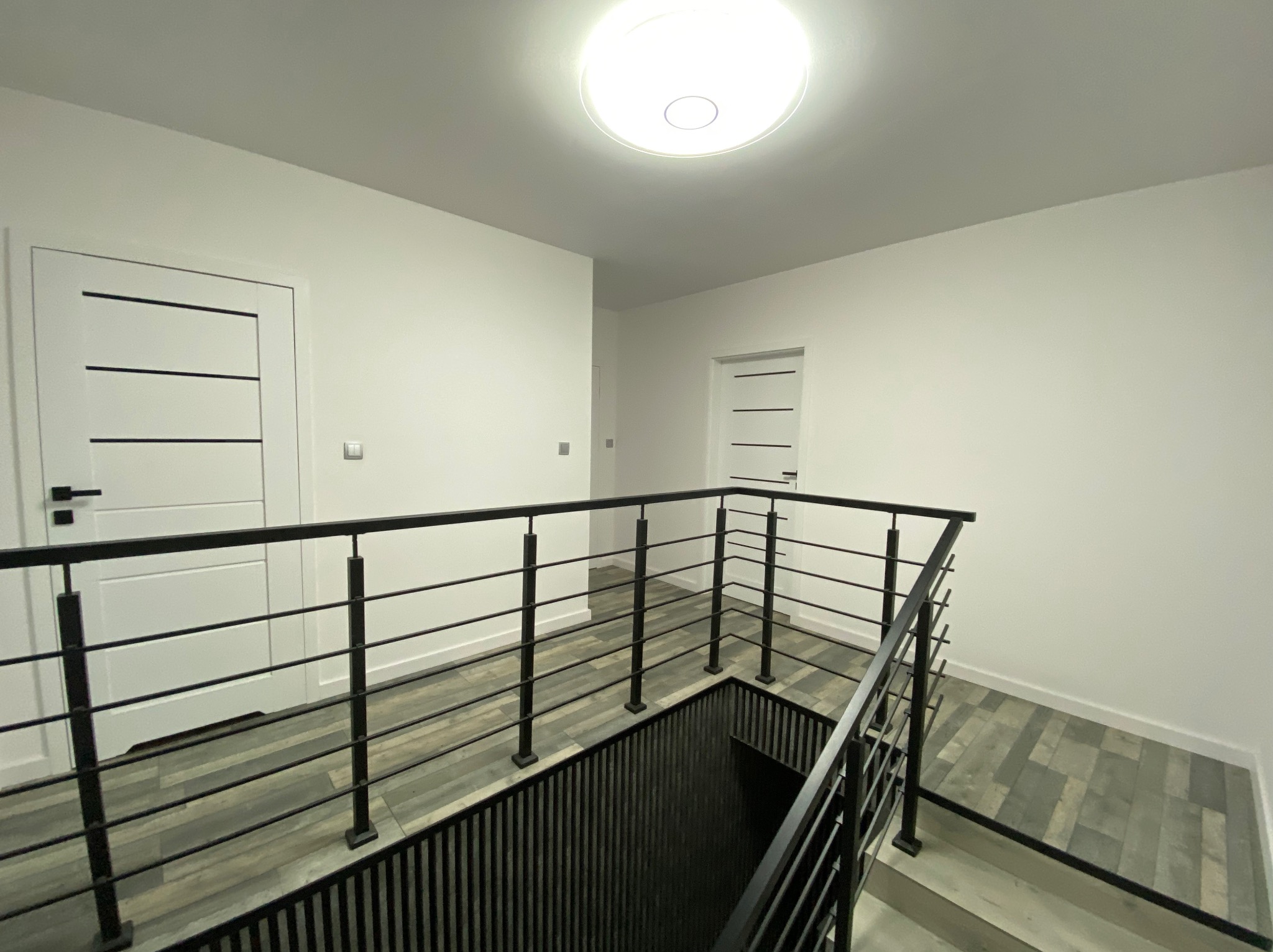 Wykończone wnętrze klatki schodowej z czarną balustradą i szarymi panelami podłogowymi. Białe ściany i drzwi z poziomymi paskami. Nowoczesne oświetlenie sufitowe.