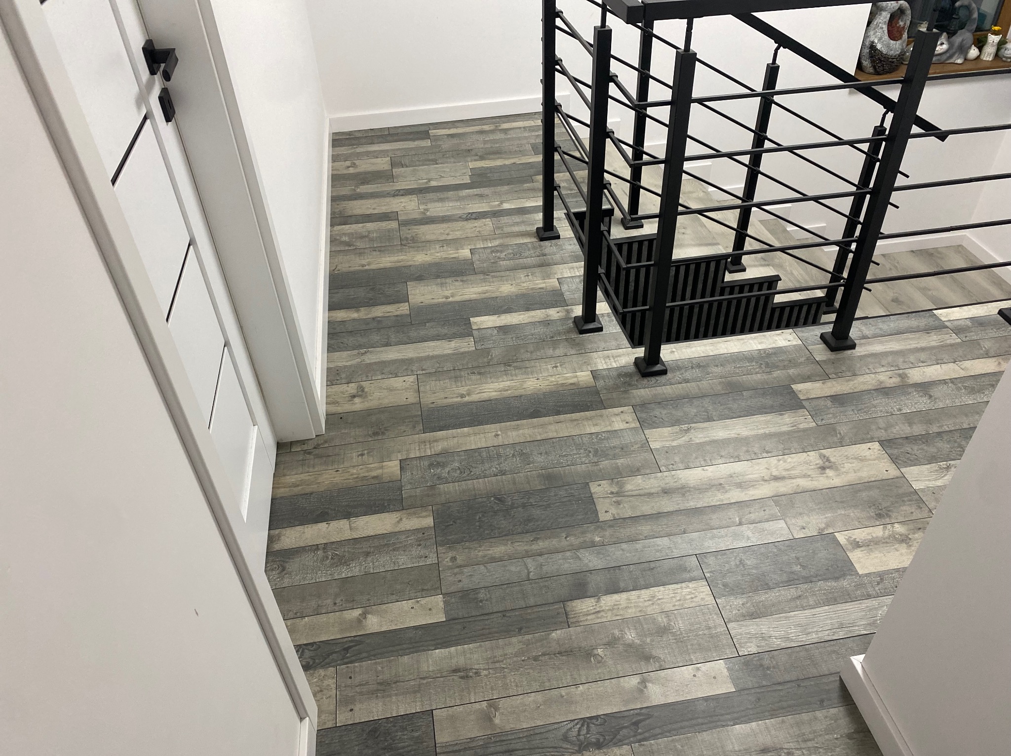 Szara podłoga z paneli imitujących drewno, ułożona w korytarzu z białą ścianą i czarną balustradą schodów. Widok z góry, z perspektywy wejścia.