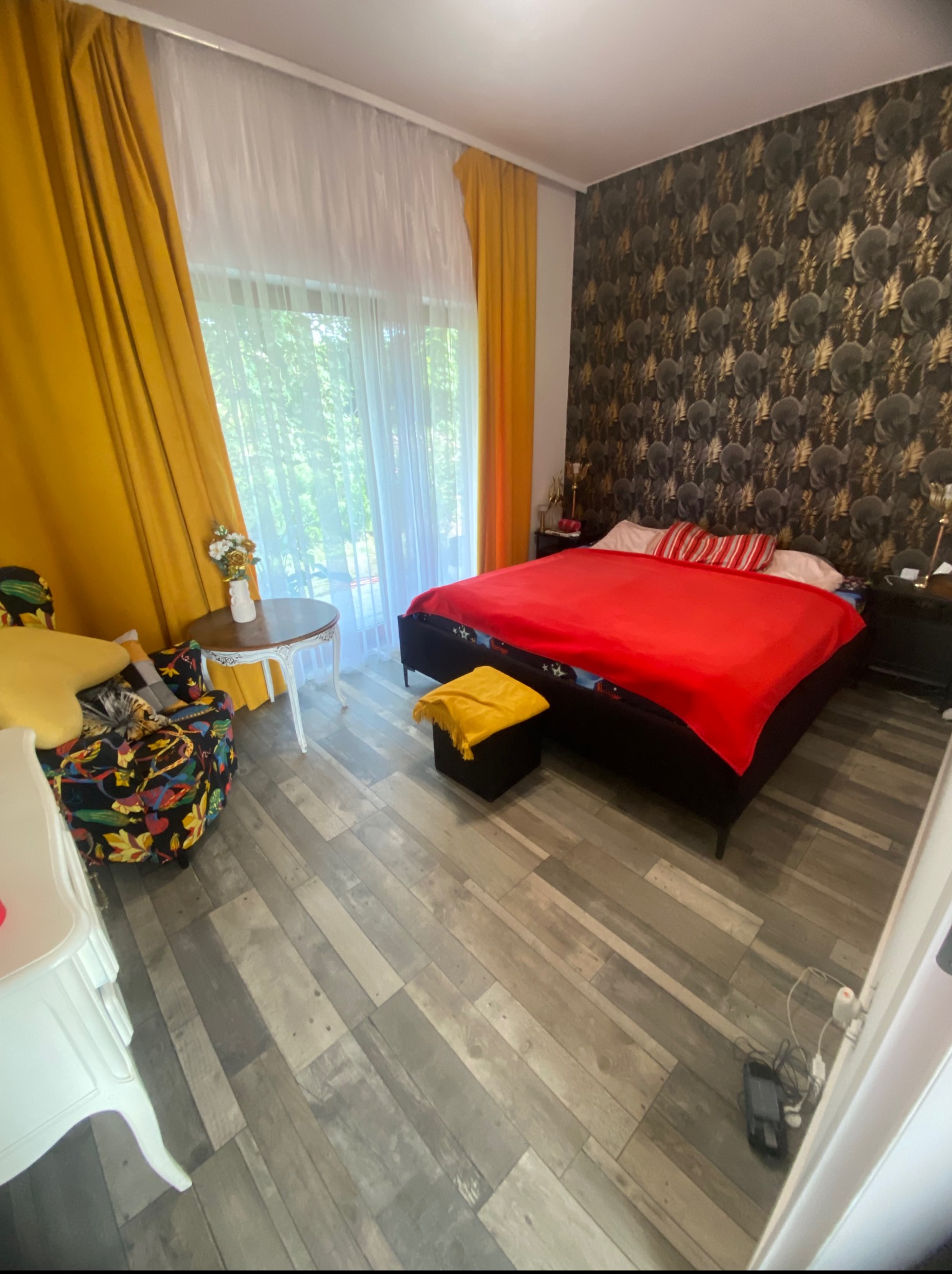 Komfortowa sypialnia z łóżkiem, wzorzystą tapetą i drewnianą podłogą. Żółte zasłony wpadają w oko, a kwiaty na stoliku dodają uroku.