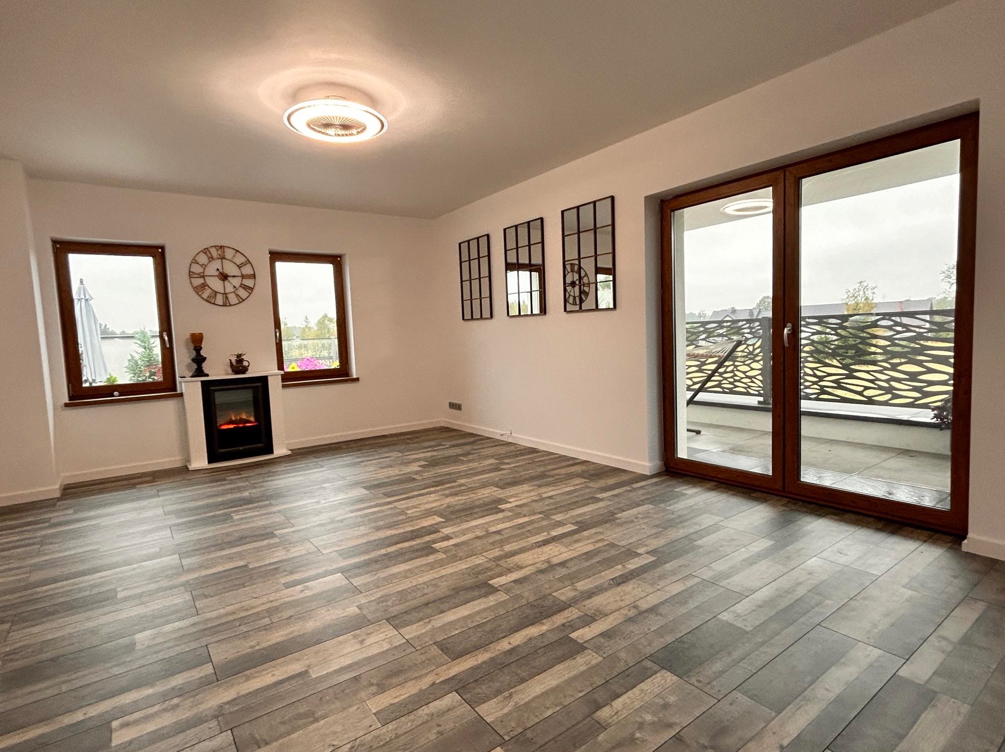 Salon z panelami podłogowymi imitującymi drewno, kominkiem elektrycznym i oknami z widokiem na zewnątrz. Dekoracyjne lustra na ścianie.