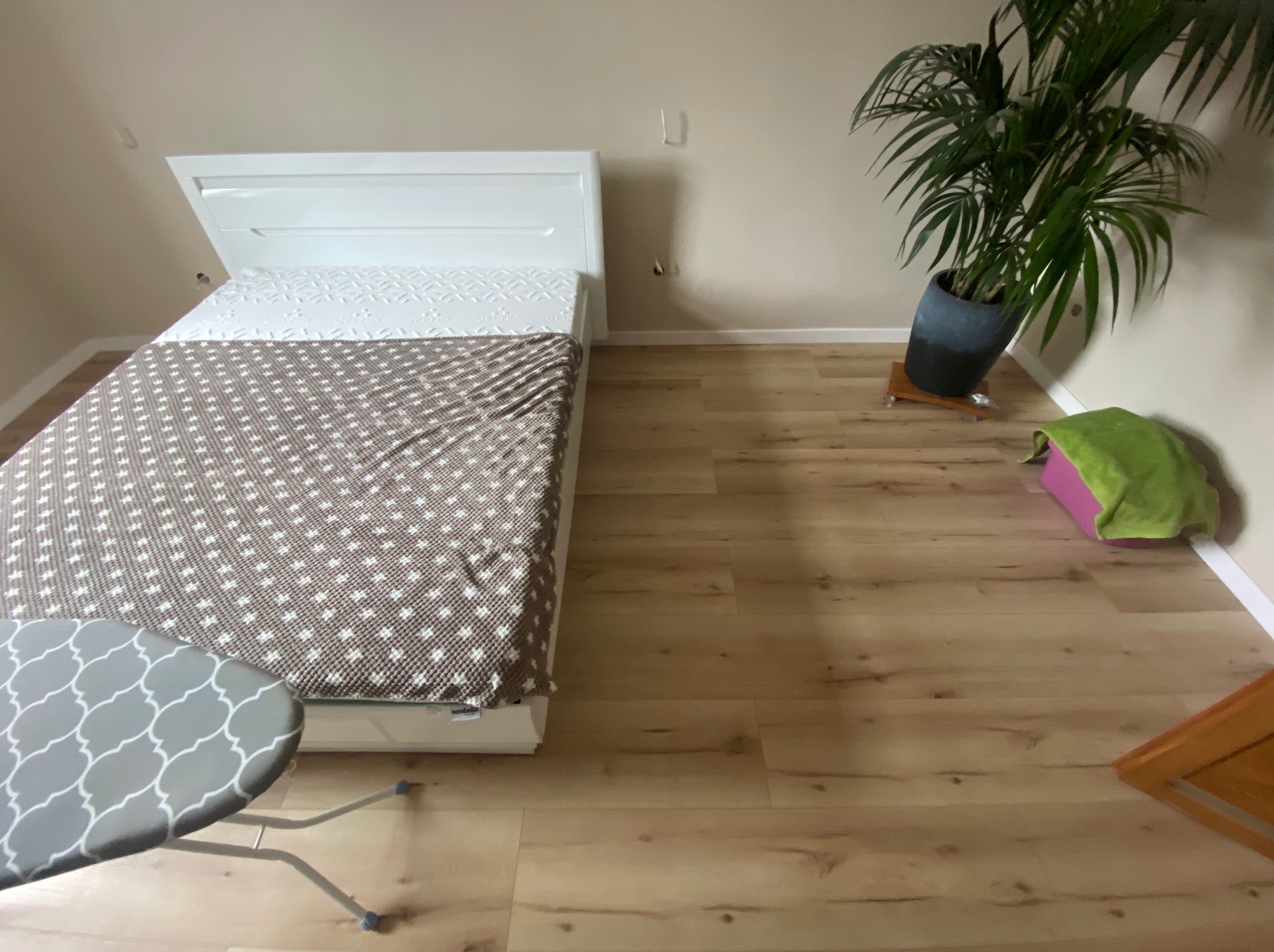 Jasne panele podłogowe w pokoju z łóżkiem, deską do prasowania i palmą w doniczce. Widok z góry na wykończone wnętrze.