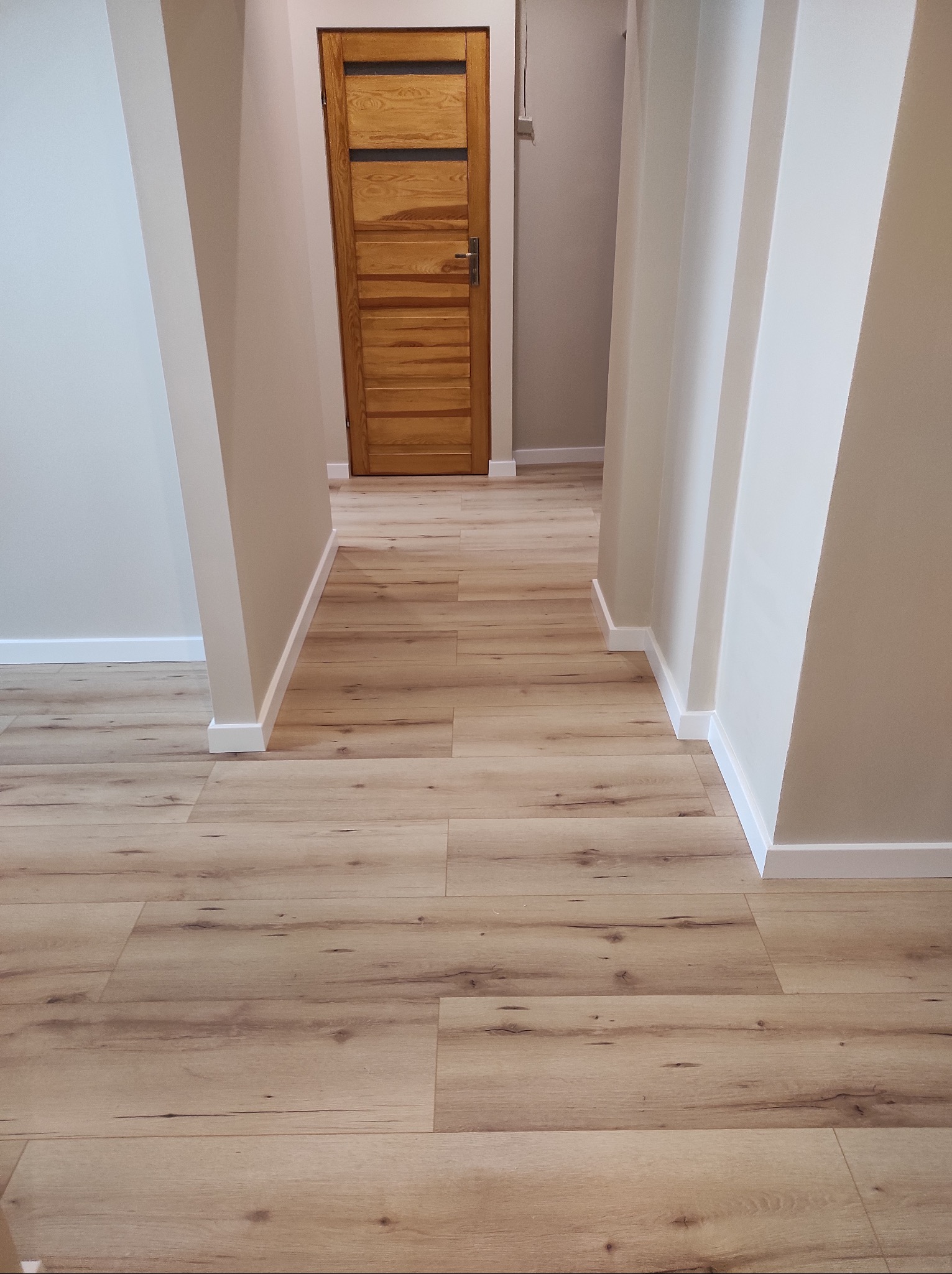 Nowoczesny korytarz z panelami podłogowymi w odcieniach drewna i drewnianymi drzwiami w tle. Ściany w jasnych, neutralnych barwach.