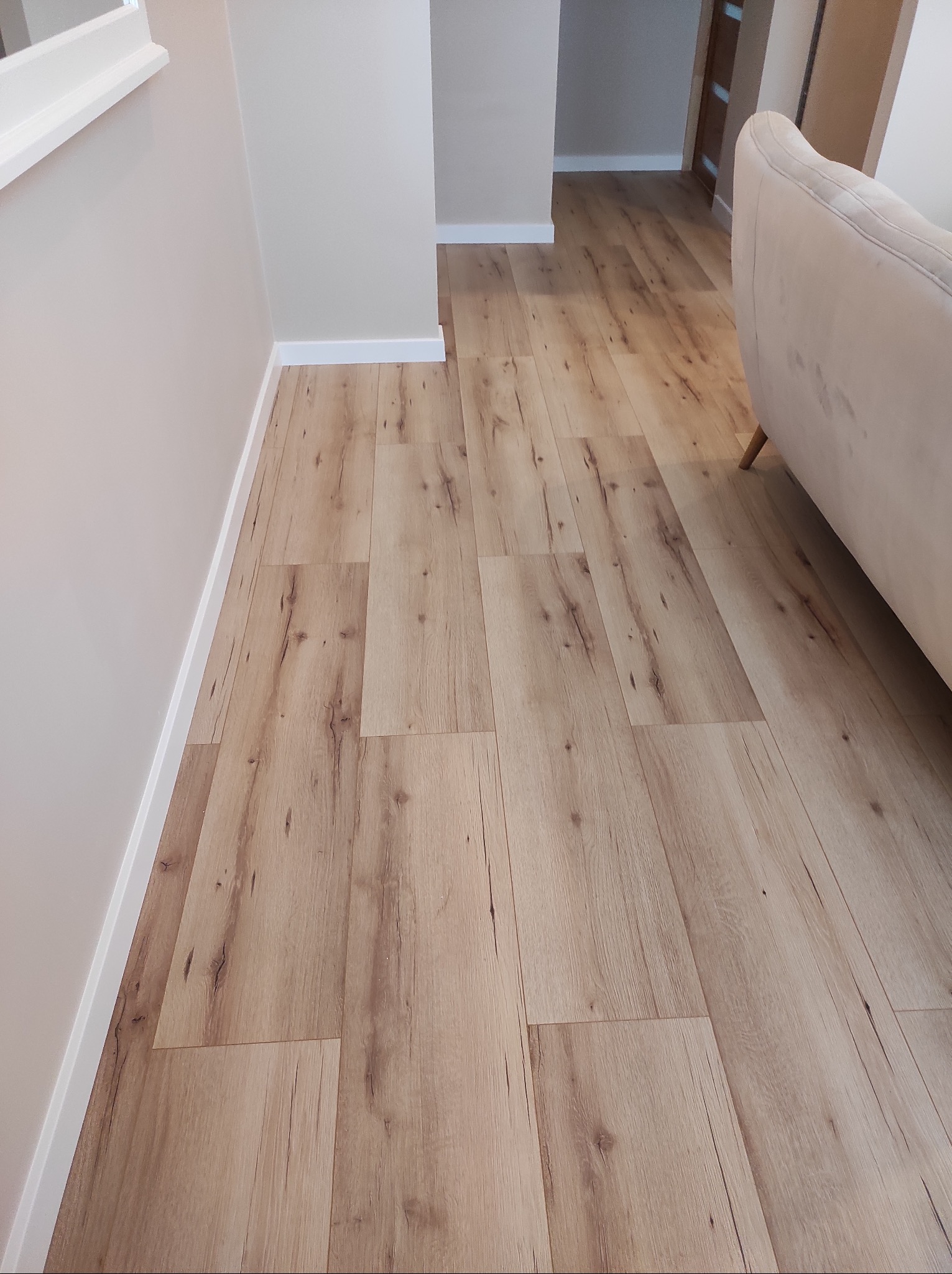 Ułożone panele podłogowe w odcieniach beżu, z widoczną fakturą drewna, w jasnym korytarzu. Fragment sofy w tle. Detal wykończenia przy ścianie.