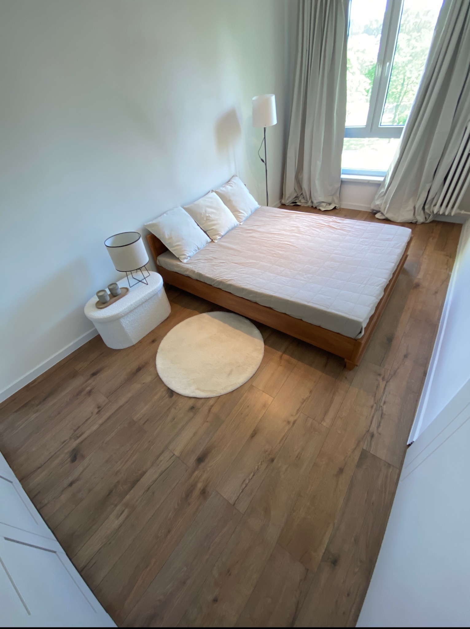 Jasna sypialnia z drewnianą podłogą, łóżkiem i okrągłym dywanem. Widok z góry ukazuje minimalistyczny design i stonowaną kolorystykę wnętrza.