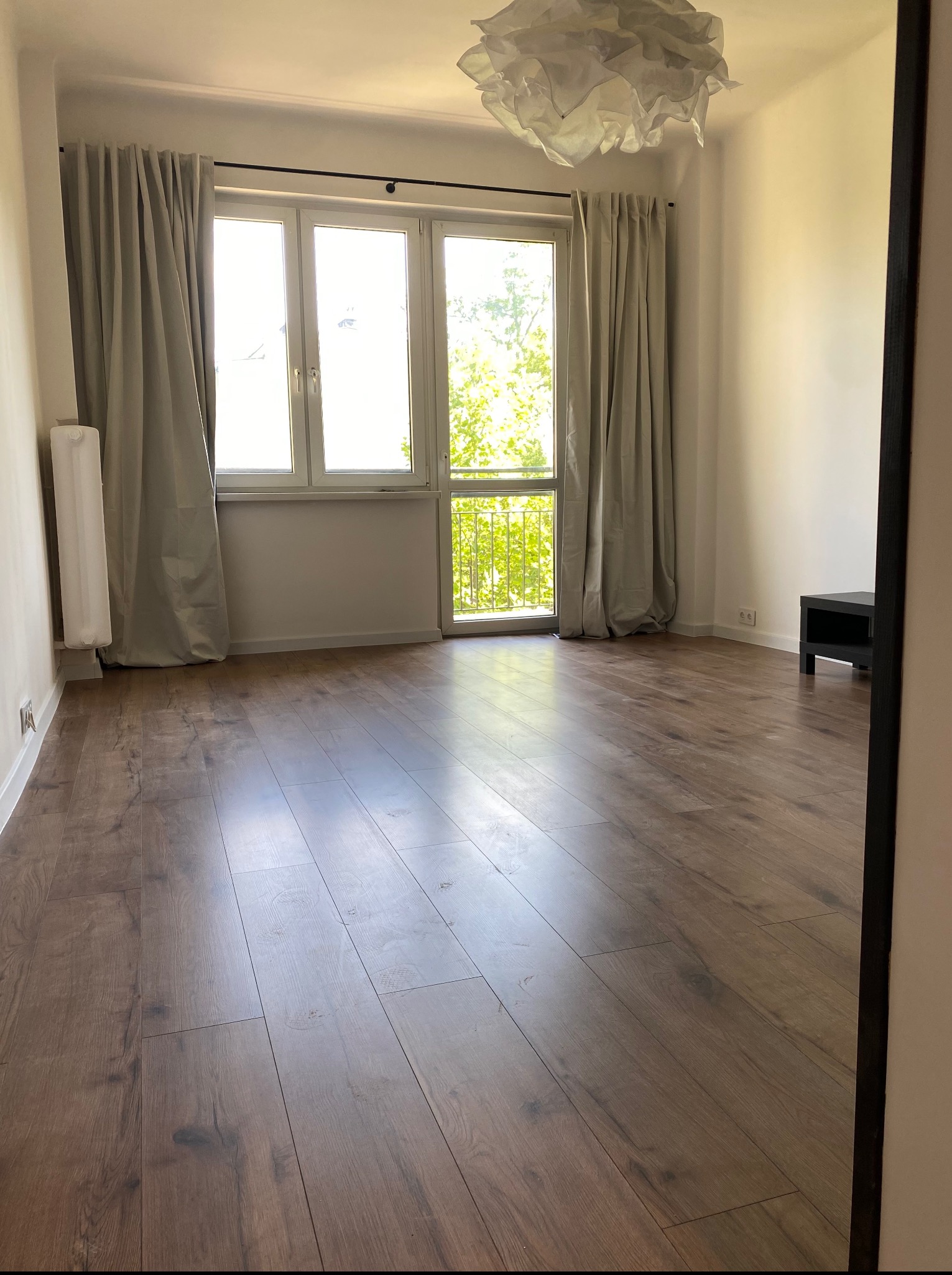 Jasne, puste wnętrze z drewnianą podłogą, dużym oknem balkonowym z widokiem na zieleń i ozdobną lampą. Widoczne szare zasłony i białe ściany.