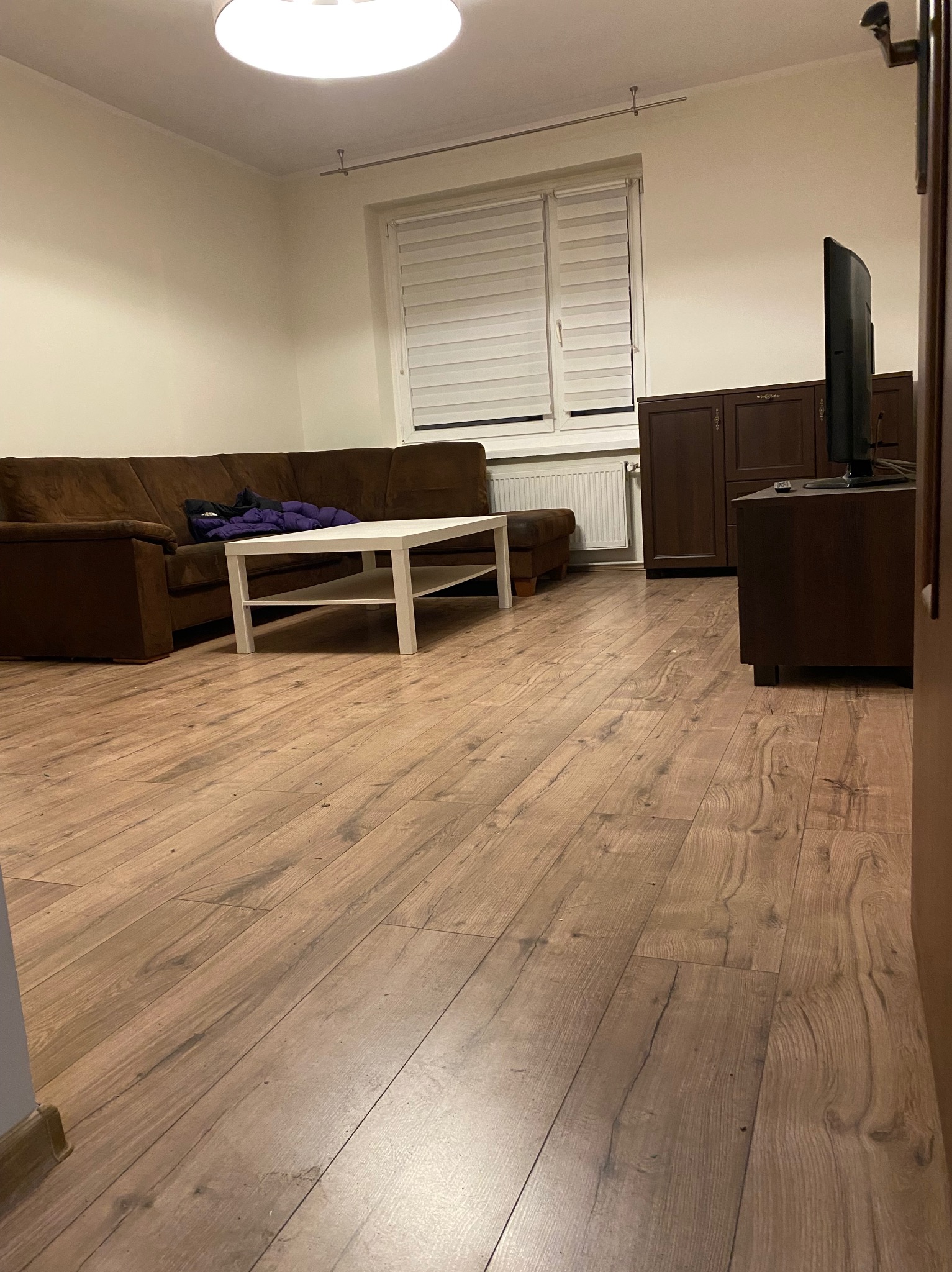 Salon z brązową sofą, białym stolikiem kawowym i parkietem w odcieniach brązu. Widoczne meble pod ścianą i okno z żaluzjami. Ciepłe oświetlenie.