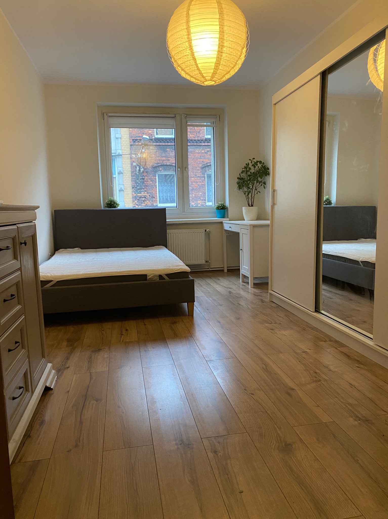 Jasne wnętrze sypialni z drewnianą podłogą, łóżkiem z szarym zagłówkiem, białą szafą z lustrem i biurkiem przy oknie z widokiem na ceglany budynek.