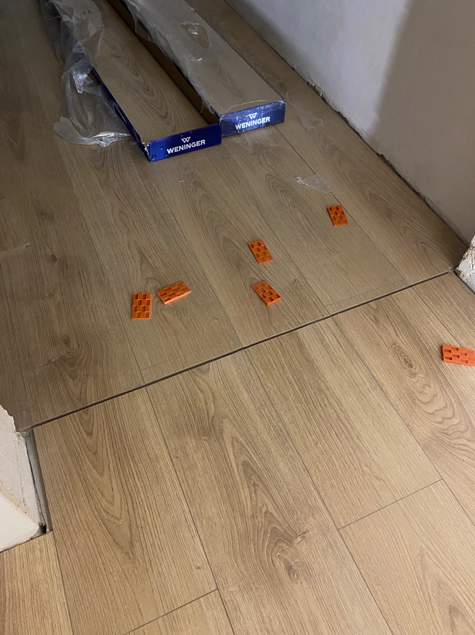 Układanie paneli podłogowych w trakcie pracy. Widoczne panele Wenninger, pomarańczowe kliny dystansowe i szczelina dylatacyjna przy ścianie.