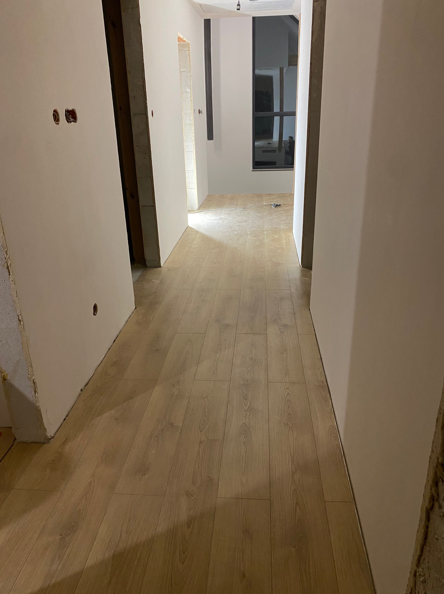 Korytarz w trakcie wykańczania: świeżo ułożone panele podłogowe, surowe ściany przygotowane do malowania, widoczne puszki elektryczne. W tle okno.