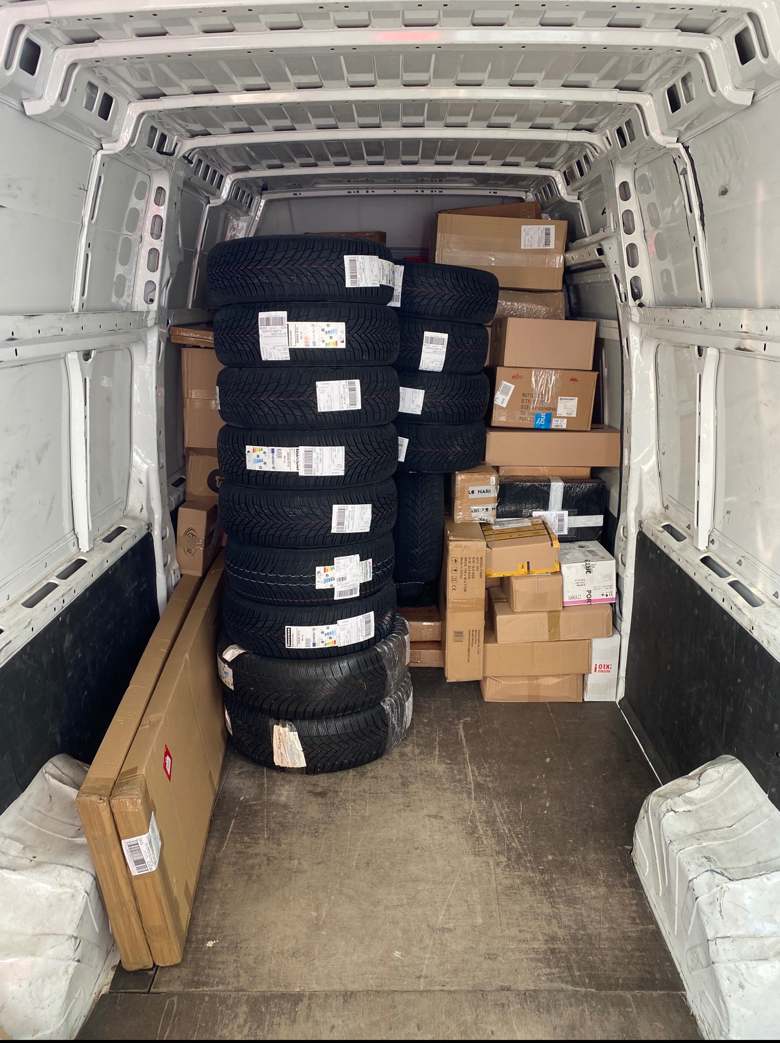 Wnętrze furgonetki wypełnionej kartonami, oponami i długimi, płaskimi paczkami, przygotowanej do transportu. Widok z tyłu pojazdu, centralne ujęcie.
