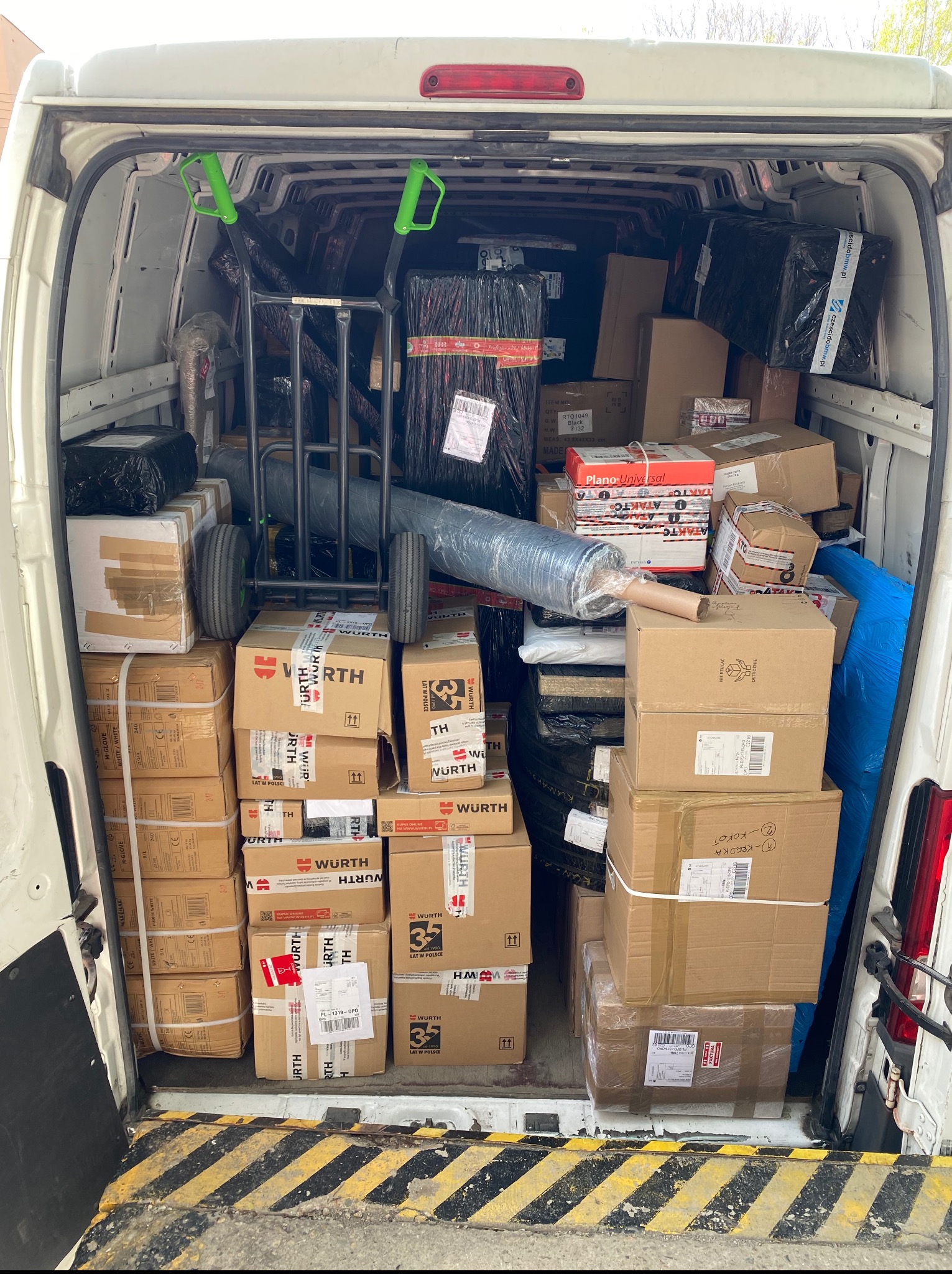 Wnętrze białego vana załadowane kartonami, wózkiem transportowym i folią stretch. Część paczek oznaczona logo Wurth. Przeprowadzka lub transport towarów.