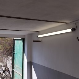 Paw-Bud-Trans Paweł Górski - Wnętrze garażu po malowaniu ścian na biało i szaro. Widoczna lampa, farby i otwarte drzwi na zewnątrz. Świeżo pomalowane ściany w garażu.