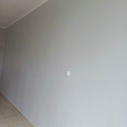 Paw-Bud-Trans Paweł Górski - Ściana pomalowana na szaro z widocznym gniazdkiem elektrycznym i fragmentem podłogi z płytek. Widoczne niedoskonałości faktury ściany.