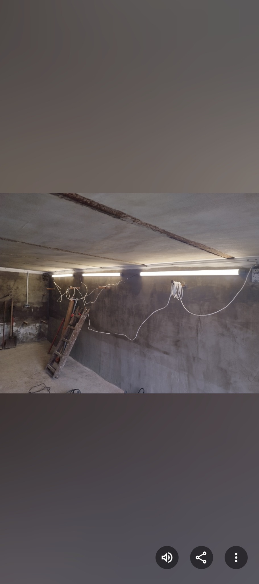 Surowy stan ścian w pomieszczeniu przed wykończeniem. Widoczne przewody, drabina i narzędzia. Ściany pokryte szarym tynkiem, oświetlenie jarzeniowe.