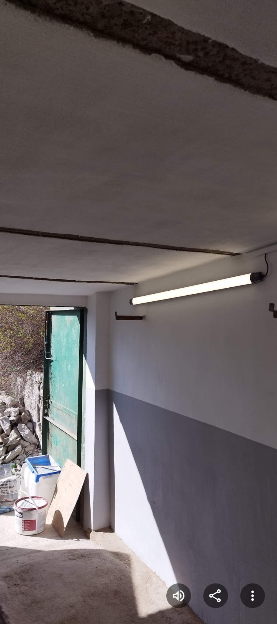 Wnętrze garażu po malowaniu ścian na biało i szaro. Widoczna lampa, farby i otwarte drzwi na zewnątrz. Świeżo pomalowane ściany w garażu.