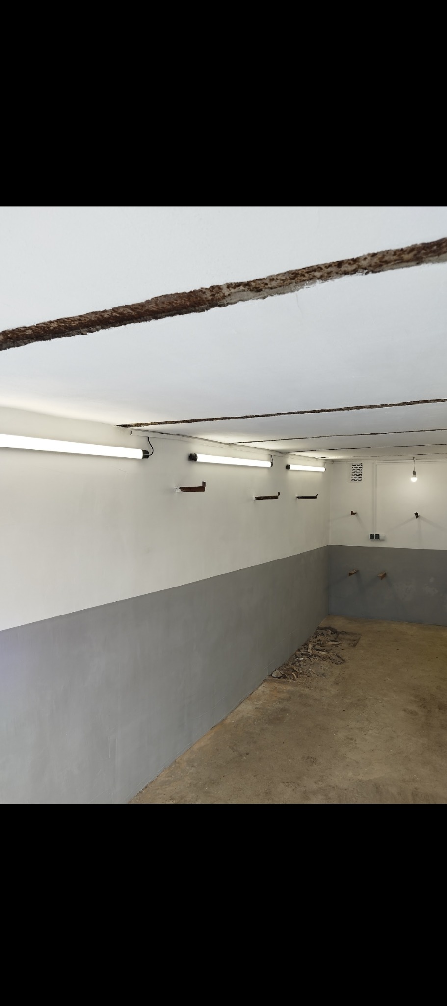 Wnętrze piwnicy po malowaniu: szara lamperia, białe ściany powyżej, widoczne lampy fluorescencyjne i ślady po instalacjach na ścianach.