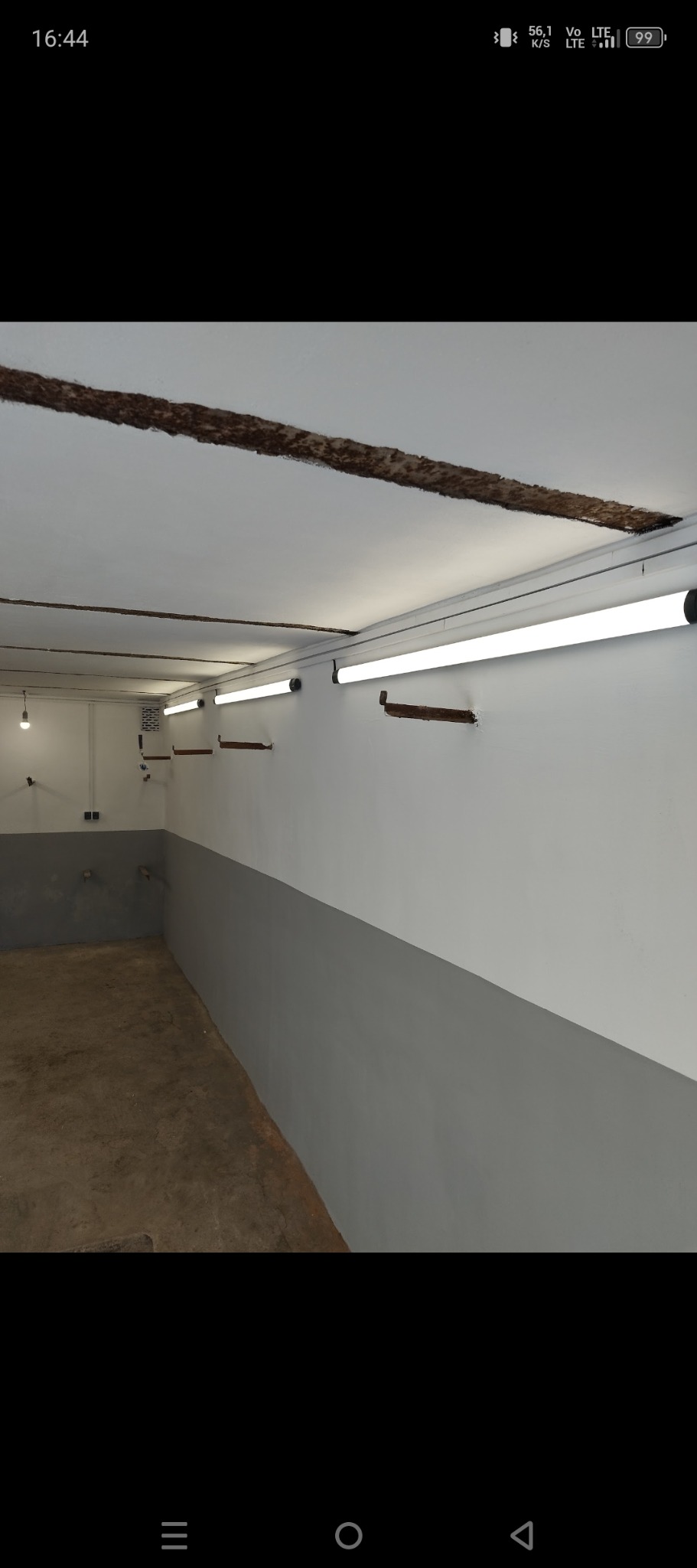 Pomieszczenie po malowaniu: biała góra, szary dół ściany, surowy fragment sufitu i lampy jarzeniowe. Widok na odnowione wnętrze z elementami do wykończenia.