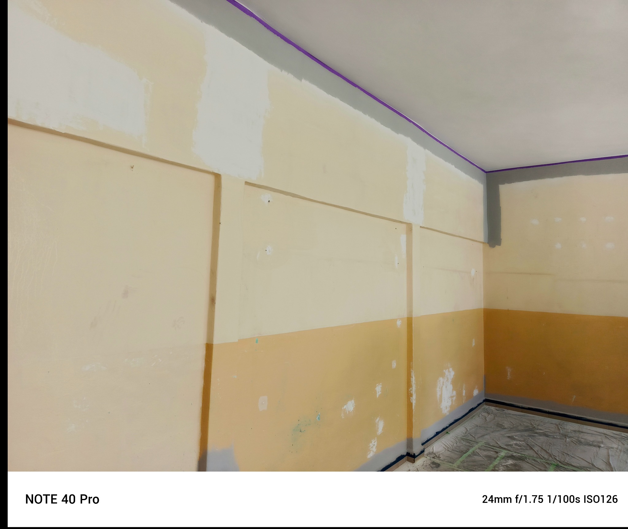 Wnętrze w trakcie prac malarskich: ściana z częściowo nałożoną białą farbą, zabezpieczone listwy przypodłogowe i sufit taśmą malarską. Widoczne ślady po szpachlowaniu.