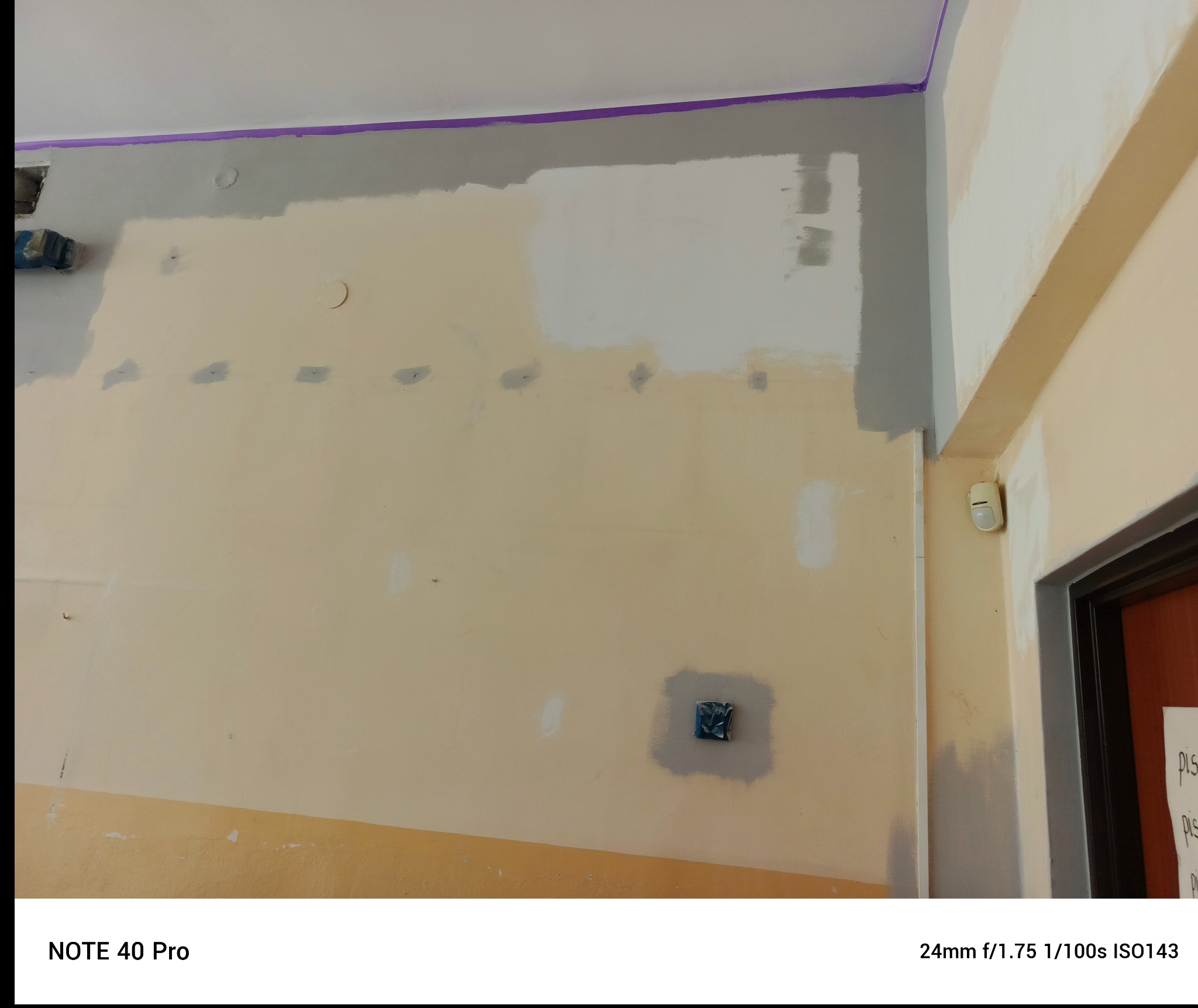 Ściana w trakcie szpachlowania i malowania z widocznymi łatami i taśmą malarską przy suficie. Fragment instalacji elektrycznej zabezpieczony folią.