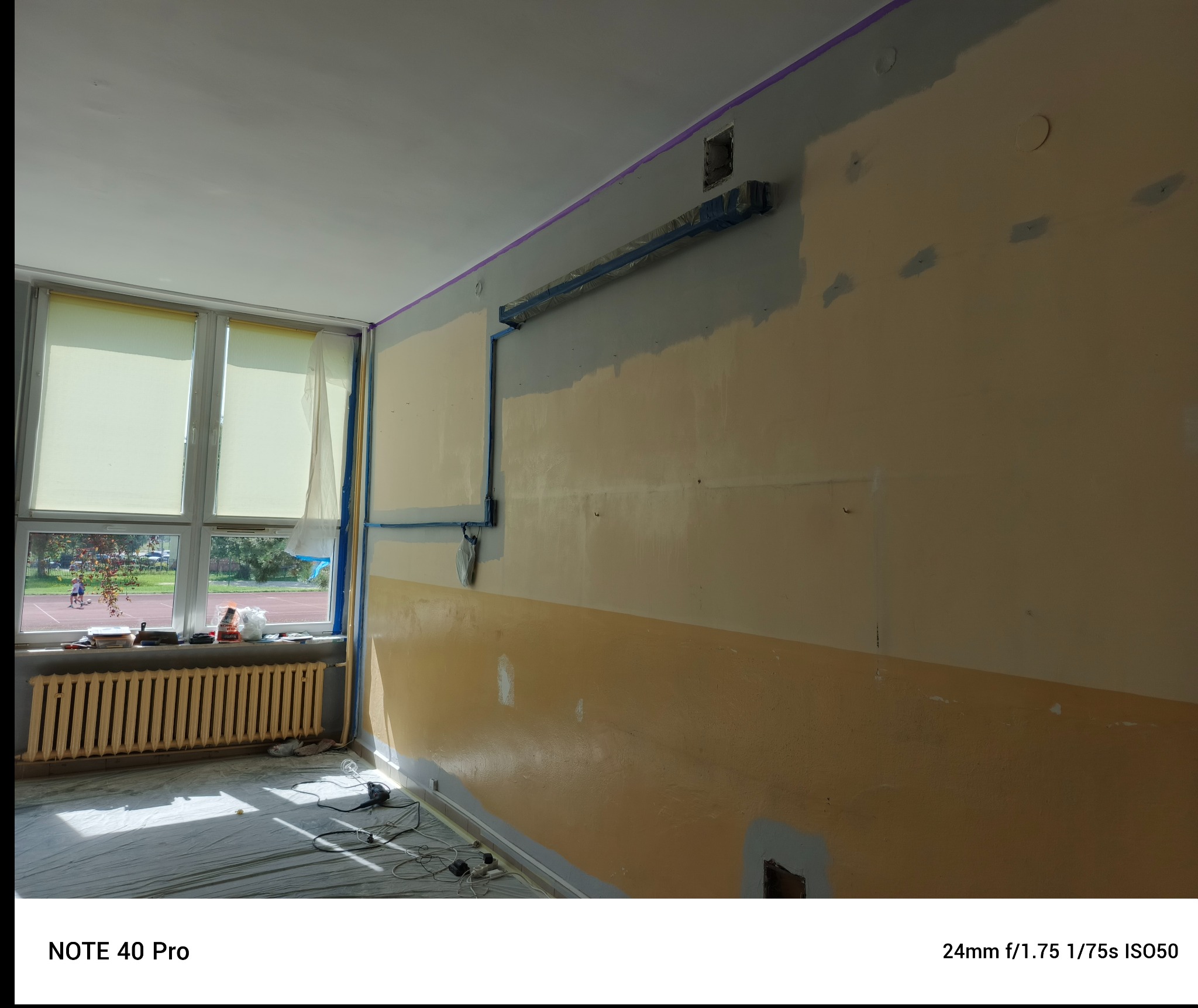 Ściana w trakcie szpachlowania, z widocznymi łatami szpachli i zabezpieczeniami okien oraz grzejnika. Przygotowanie do malowania w pomieszczeniu z oknem na zewnątrz.