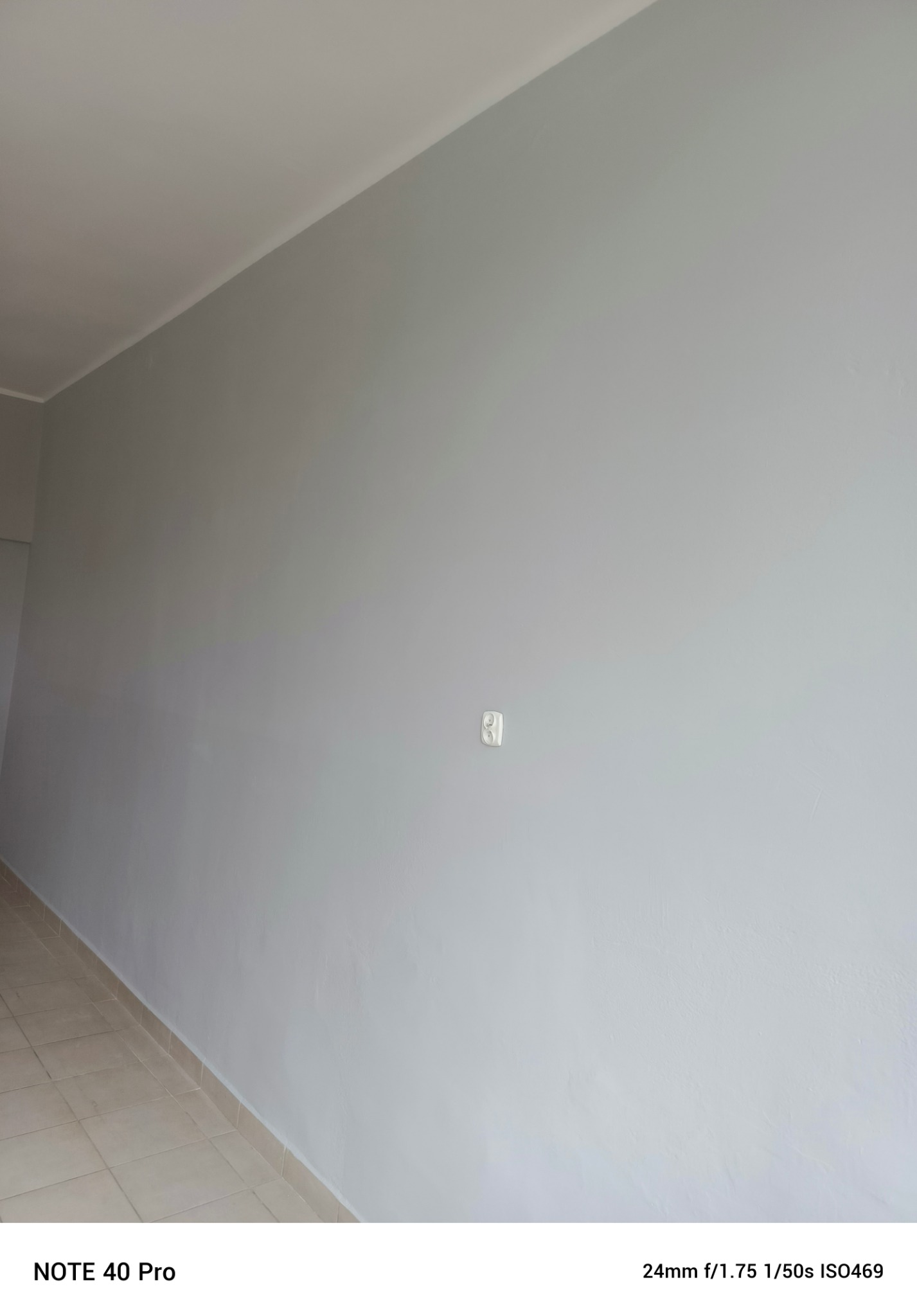 Ściana pomalowana na szaro z widocznym gniazdkiem elektrycznym i fragmentem podłogi z płytek. Widoczne niedoskonałości faktury ściany.