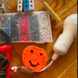 Narzędzia elektryczne: miernik, złączki, gniazda, listwa zaciskowa i żarówka LED na parkiecie. Pomarańczowy uchwyt z uśmiechem dodaje unikalności.