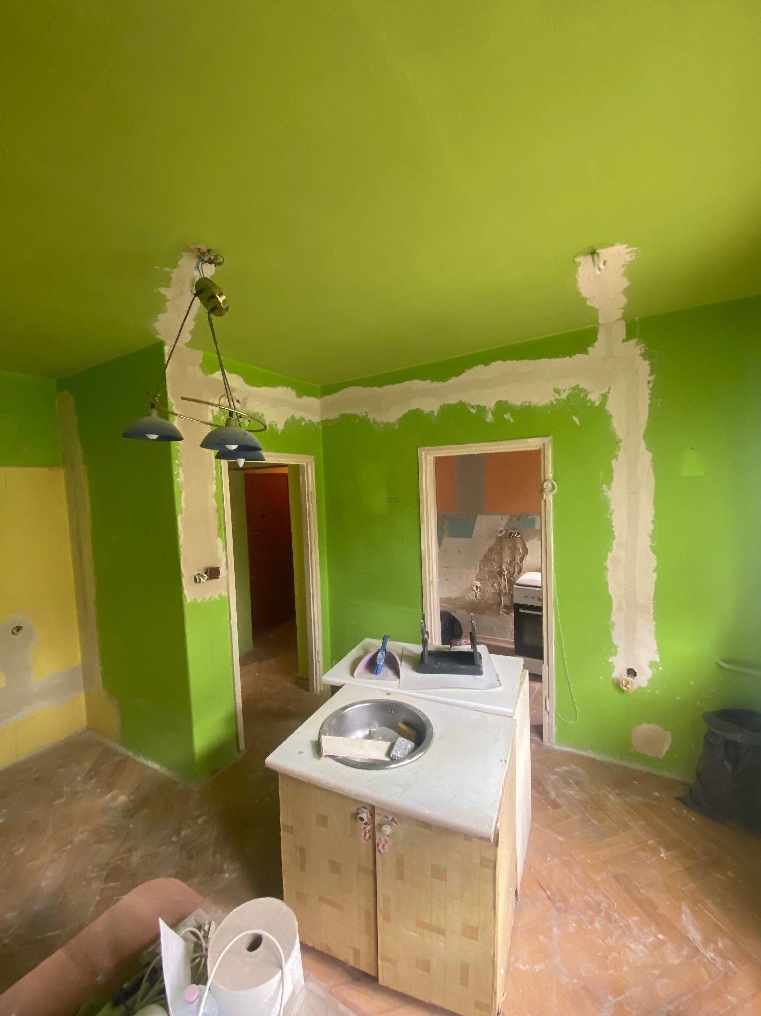 Remont kuchni w jaskrawych kolorach: zielone ściany z białymi śladami po instalacji elektrycznej, wisząca lampa i fragmenty żółtej ściany. Widoczne ślady prac remontowych.
