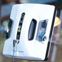 Robot do mycia okien Hobot przytwierdzony do szyby. Widoczny przewód zasilający i zbiornik na płyn. Tło rozmyte, zieleń i elementy architektoniczne.