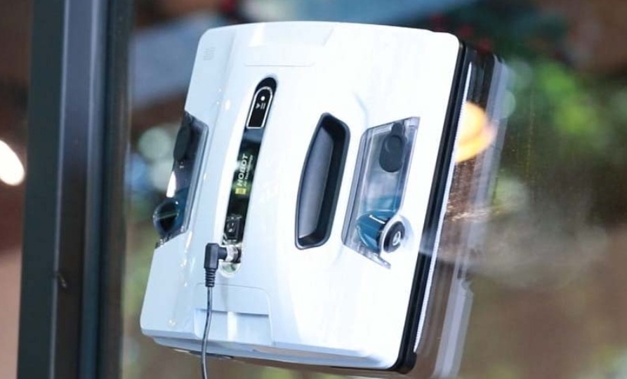 Robot do mycia okien Hobot przytwierdzony do szyby. Widoczny przewód zasilający i zbiornik na płyn. Tło rozmyte, zieleń i elementy architektoniczne.