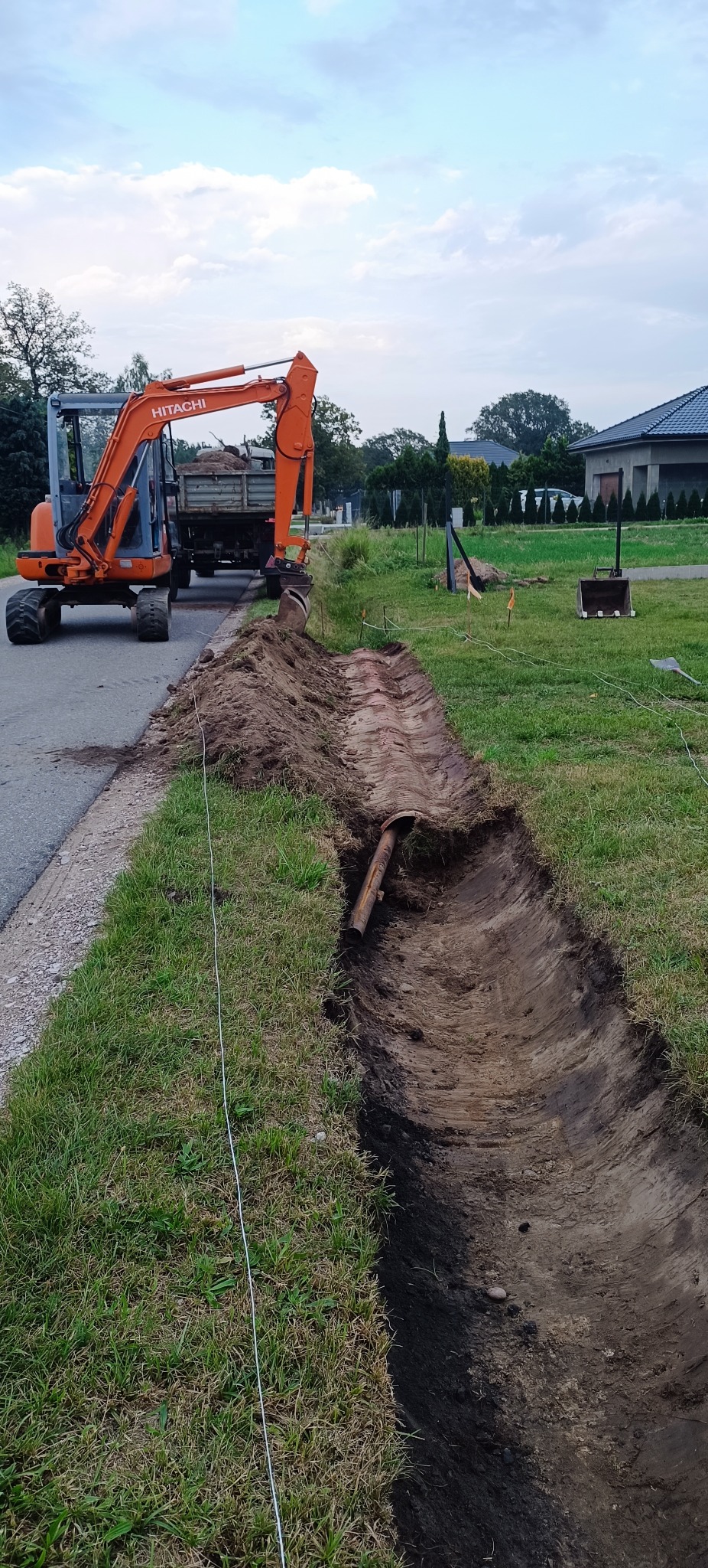 Wykopany rów pod przyłącze, widoczna rura w ziemi, pomarańczowa koparka Hitachi i wywrotka w tle. Prace ziemne na trawniku w pobliżu drogi.