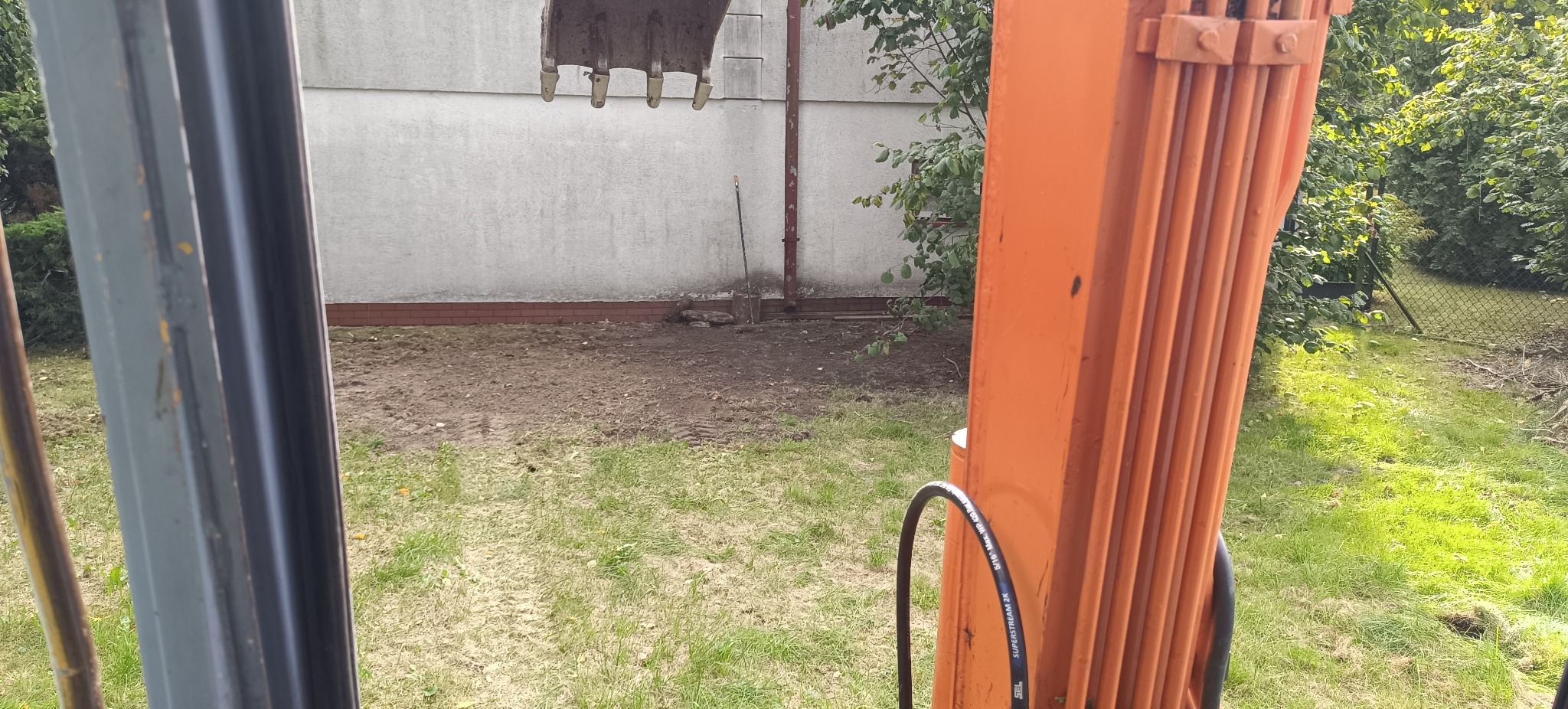 Prace ziemne w Pabianicach: łyżka koparki nad przygotowanym terenem pod instalacje, widoczna łopata. Wykopy pod szambo lub oczyszczalnię.