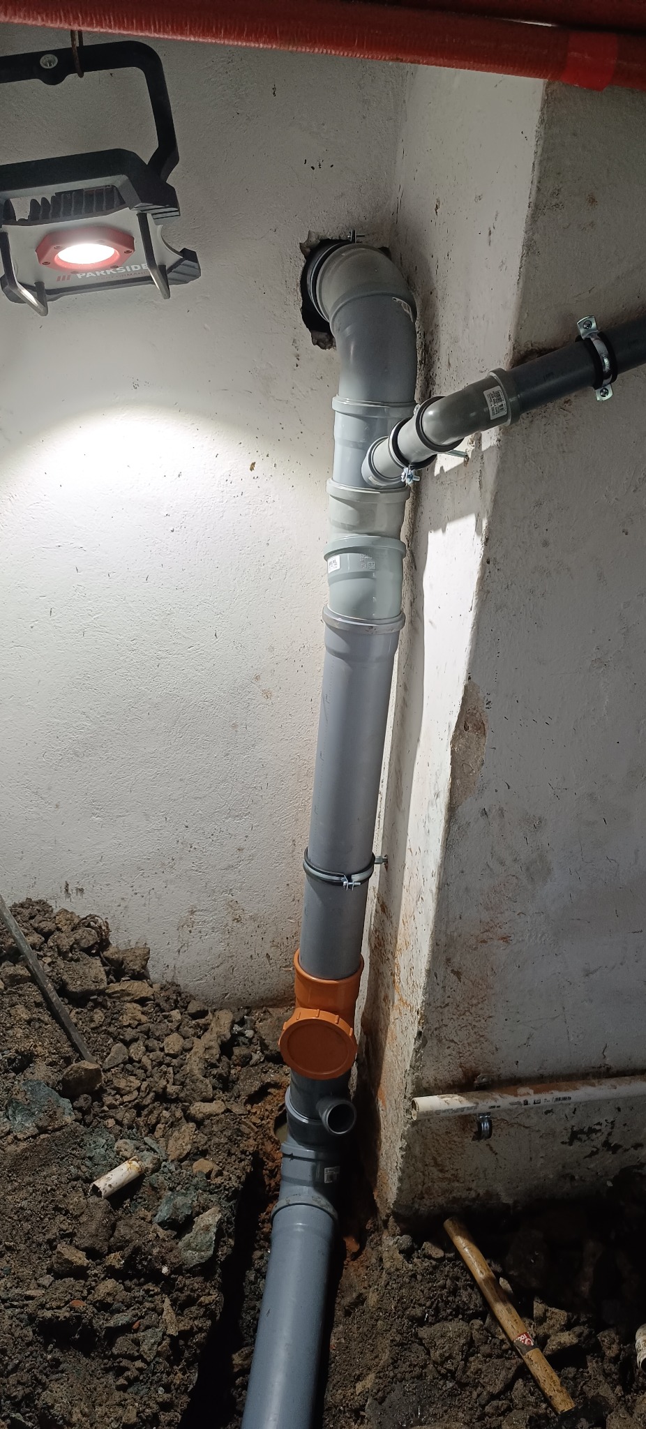 Szara instalacja kanalizacyjna z pomarańczowym elementem, zamontowana przy ścianie z oświetleniem roboczym na tle ziemi i gruzu. Widoczne złącza i uchwyty mocujące.