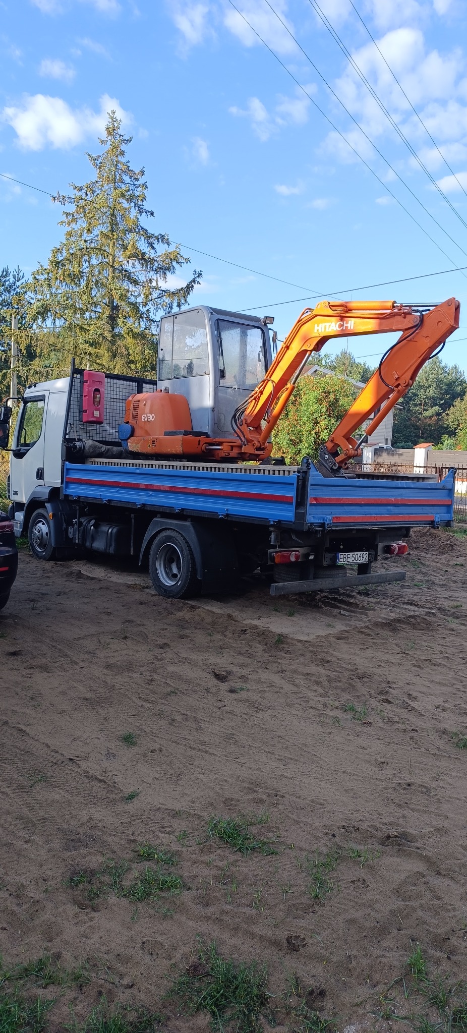 Pomarańczowa koparka Hitachi EX30 na niebieskiej platformie ciężarówki na piaszczystym podłożu, tło z zielenią i błękitnym niebem. Widoczny numer rejestracyjny.