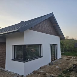 Elewacje z klasą - Nowoczesny dom jednorodzinny z białą elewacją i drewnianymi akcentami, widoczny z boku, w trakcie prac wykończeniowych na zewnątrz. Uwagę zwraca duże okno.