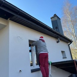 Elewacje z klasą - Pracownik w czapce Mikołaja na drabinie, uszczelnia okno w świeżo otynkowanej, białej elewacji domu z czarnym dachem i rynnami. Błękitne niebo w tle.
