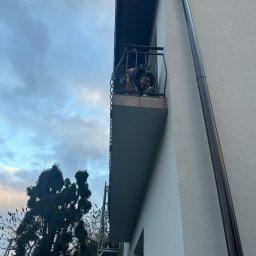 Elewacje z klasą - Osoba na balkonie budynku z jasną elewacją, widoczna z dołu. Obok budynku drzewo i rusztowanie. Ujęcie z dołu, pochmurne niebo w tle.