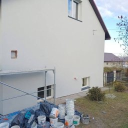 Elewacje z klasą - Świeżo pomalowana, jasnoszara elewacja domu jednorodzinnego z czerwonym dachem. Widoczne rusztowanie, puste wiadra po farbie i folia malarska na trawniku.