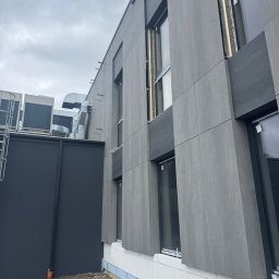 Elewacje z klasą - Szara elewacja budynku z widoczną warstwą ocieplenia przy gruncie. Widoczne okna i elementy wentylacji na dachu. Ujęcie z dołu, pochmurne niebo.