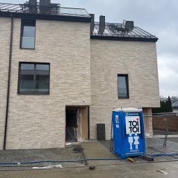 Elewacje z klasą - Nowoczesny budynek z jasną elewacją z cegły klinkierowej, widoczne okna z czarnymi ramami, fragment dachu i przenośna toaleta TOI TOI na placu budowy.